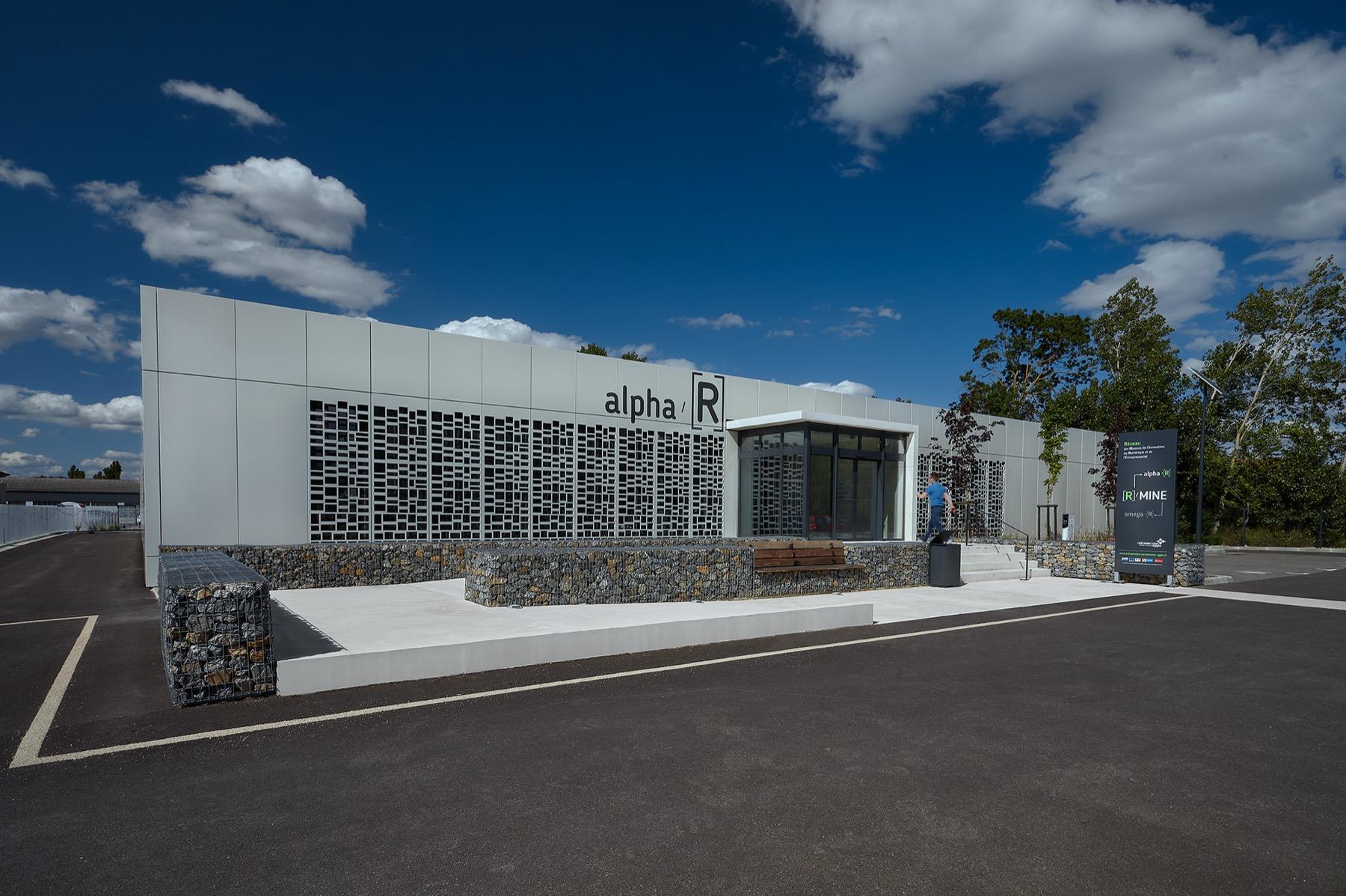 Alpha R — Façade blanche du bâtiment Alpha R aux panneaux ajourés et murets en gabions, sous un ciel bleu nuageux