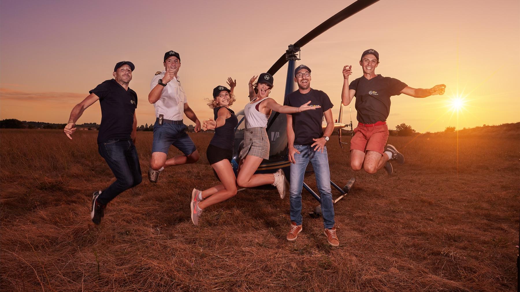 Lift Off — Équipe de six personnes sautant de joie devant un hélicoptère posé dans un champ, au coucher du soleil