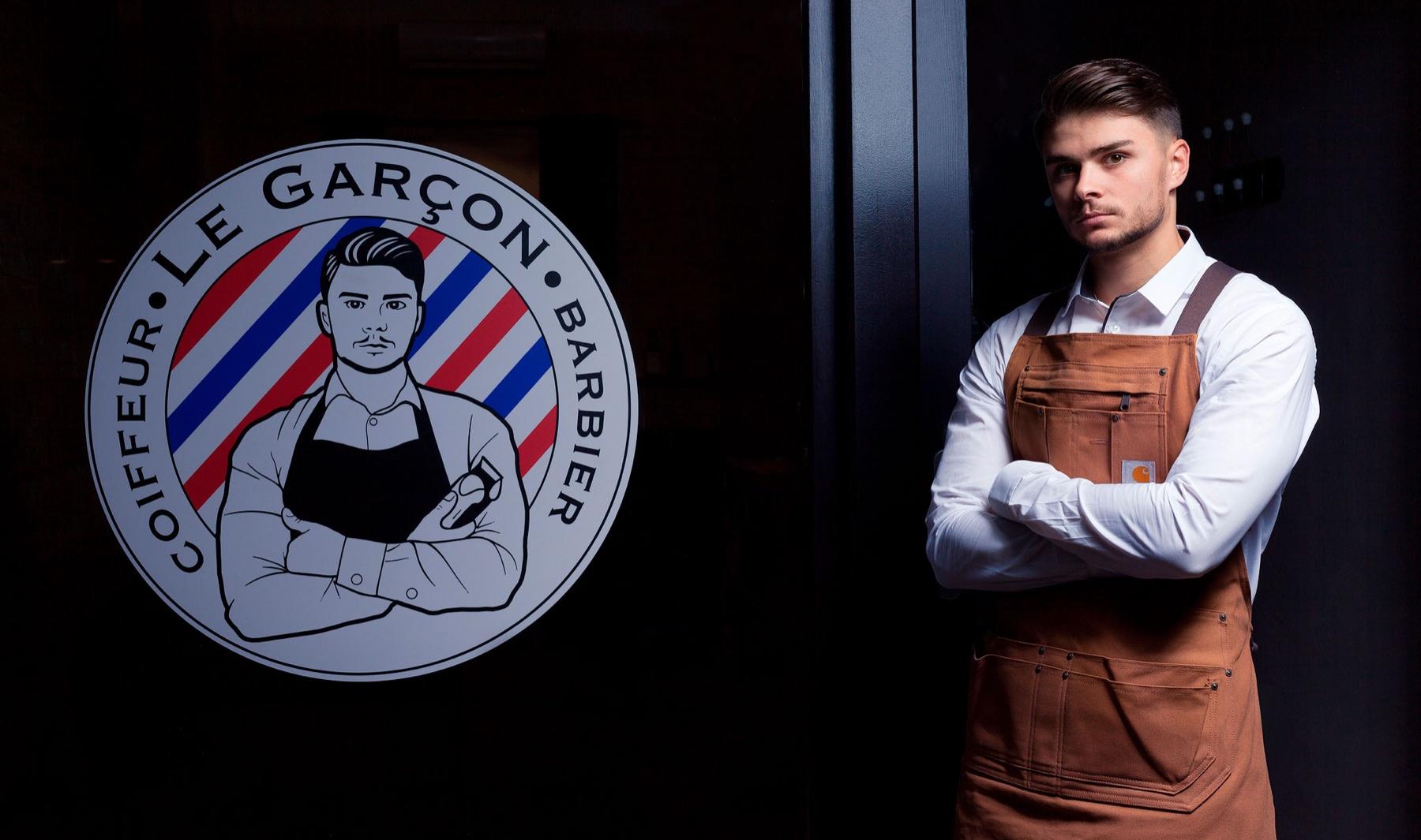 Le Garçon — Barbier en tablier, bras croisés, à côté du logo Le Garçon Barbier