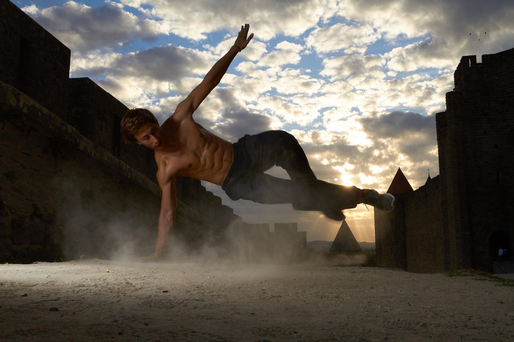 Ramparts — Pratiquant de parkour en appui sur une main, suspendu en l’air entre des remparts médiévaux, à contre-jour du soleil couchant