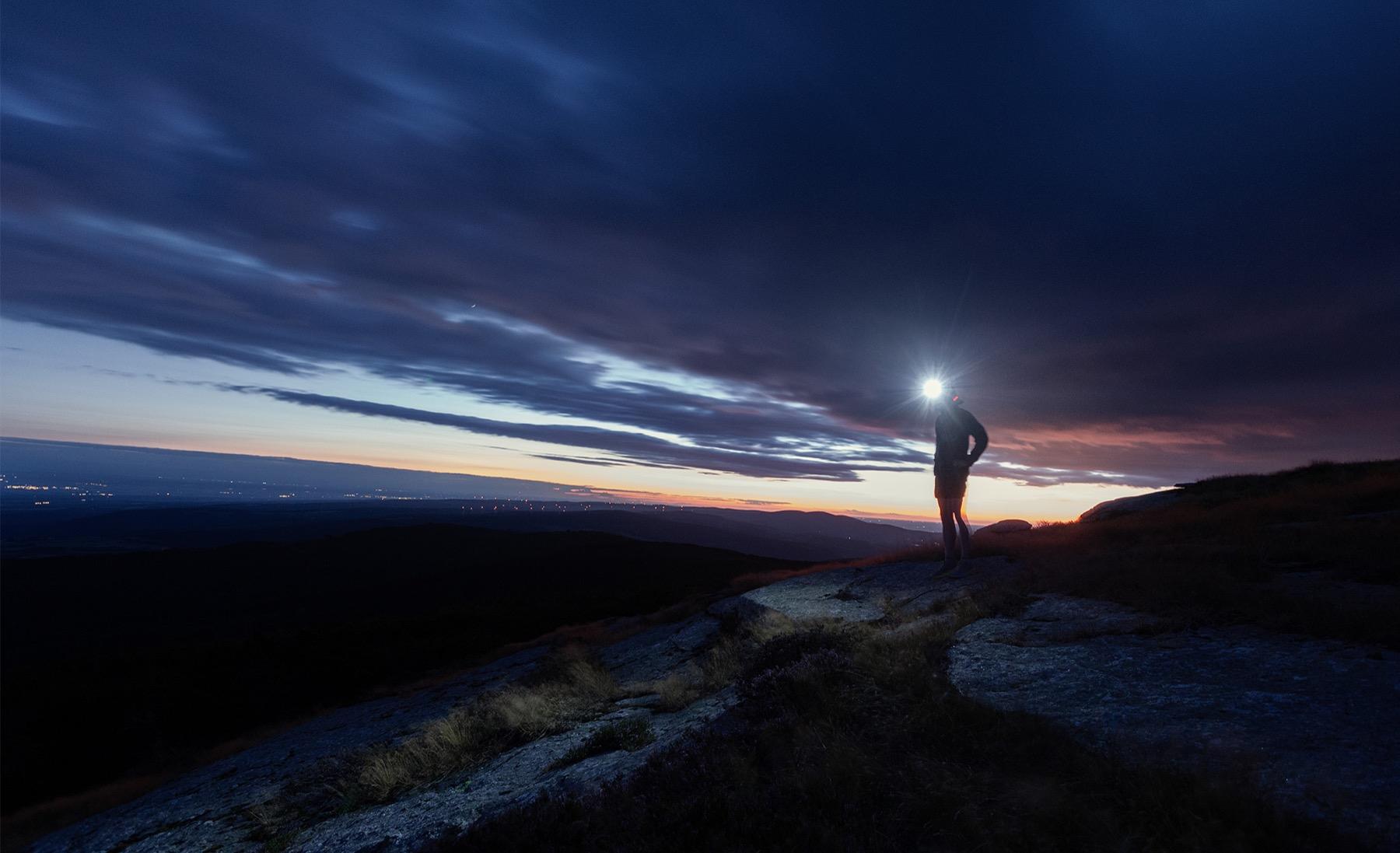 Ridge Line — Traileur à la lampe frontale sur une crête rocheuse, sous un ciel nuageux à l’heure bleue